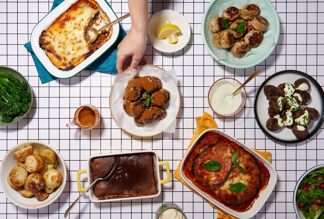Table spread of baked dishes, meatballs, and roasted vegetables with a hand reaching in.