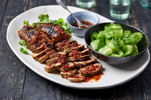 Sliced barbecued pork with chilli dipping sauce and cucumber salad on a platter.