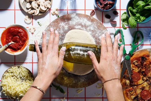 Hands rolling out pizza dough with a wine bottle surrounded by cooking ingredients.