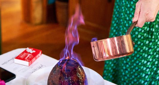 Christmas pudding being flambéed with brandy from a copper pot, blue flame rising.