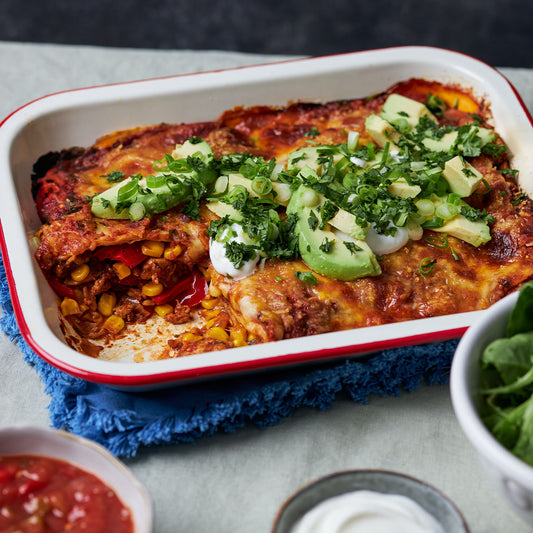 Baking dish of layered chicken enchiladas topped with melted cheese, avocado, sour cream, spring onions and coriander.
