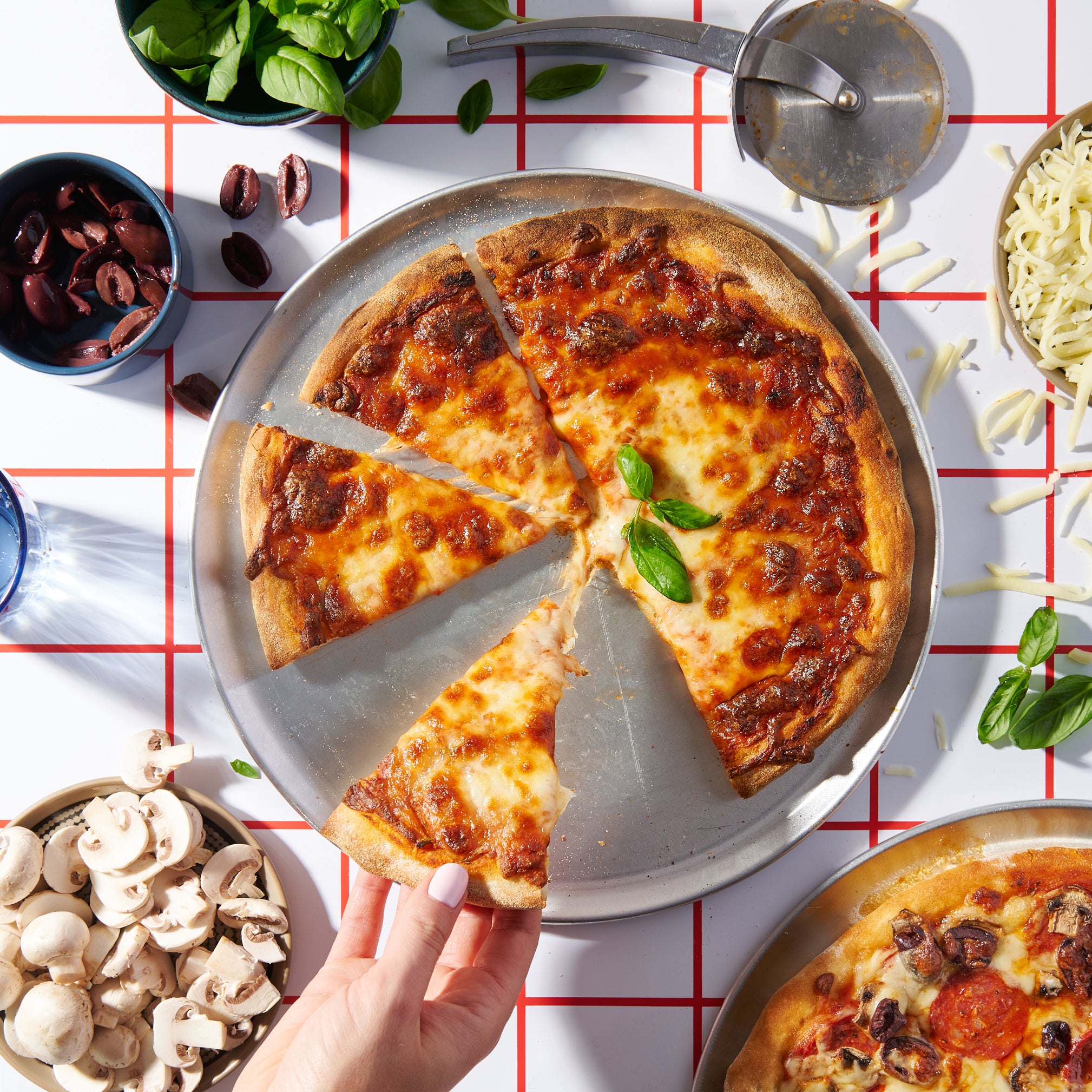 Hand reaching for a slice of Margherita pizza with golden crust and basil garnish, surrounded by bowls of olives, mushrooms, and cheese.