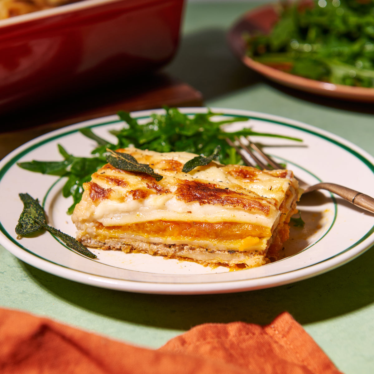 Slice of roast pumpkin and ricotta lasagne plated with crispy sage leaves and a rocket salad