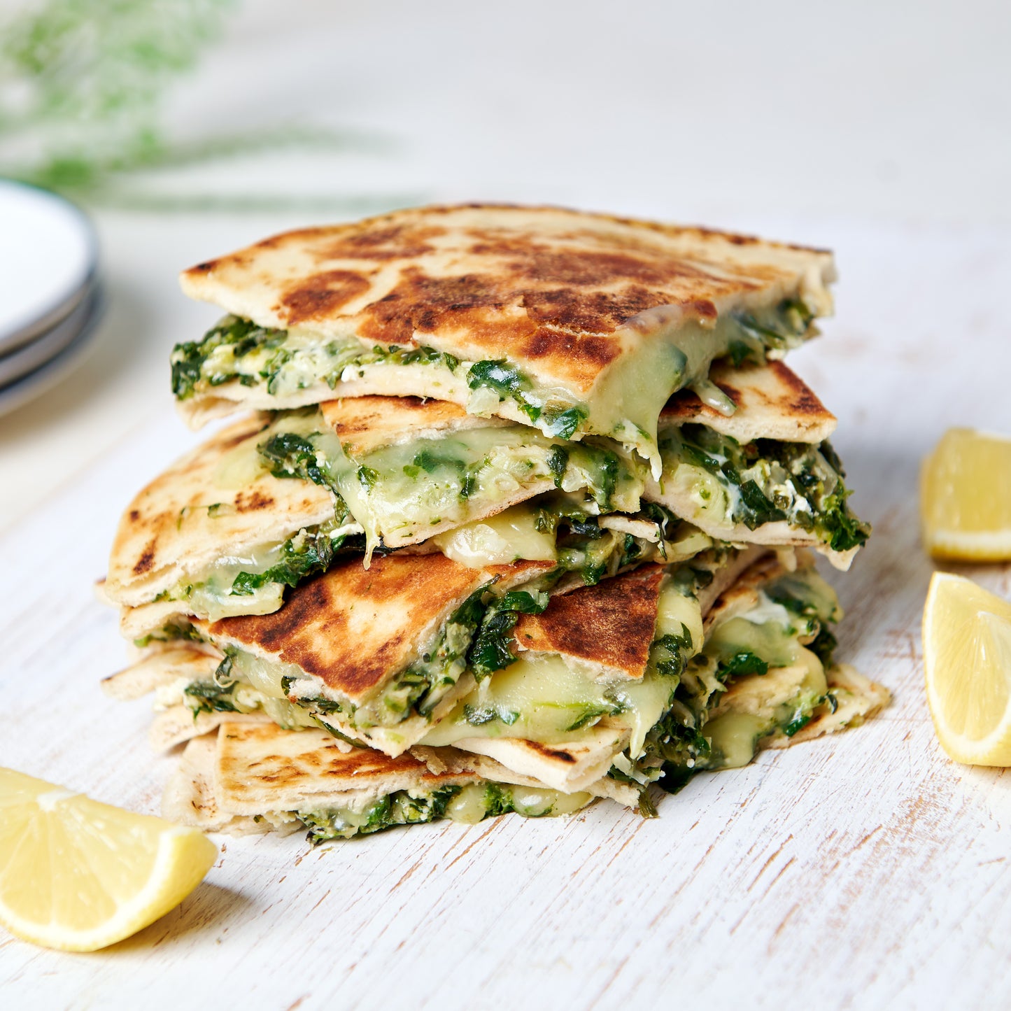 Stack of golden spinach and three cheese flatbreads, oozing with melted mozzarella, feta and ricotta, served with lemon wedges on a wooden board.