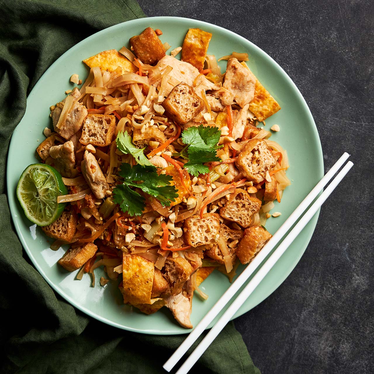 Plate of chicken pad Thai with rice noodles, tofu, peanuts, carrot and coriander, garnished with lime, served with chopsticks on the side.