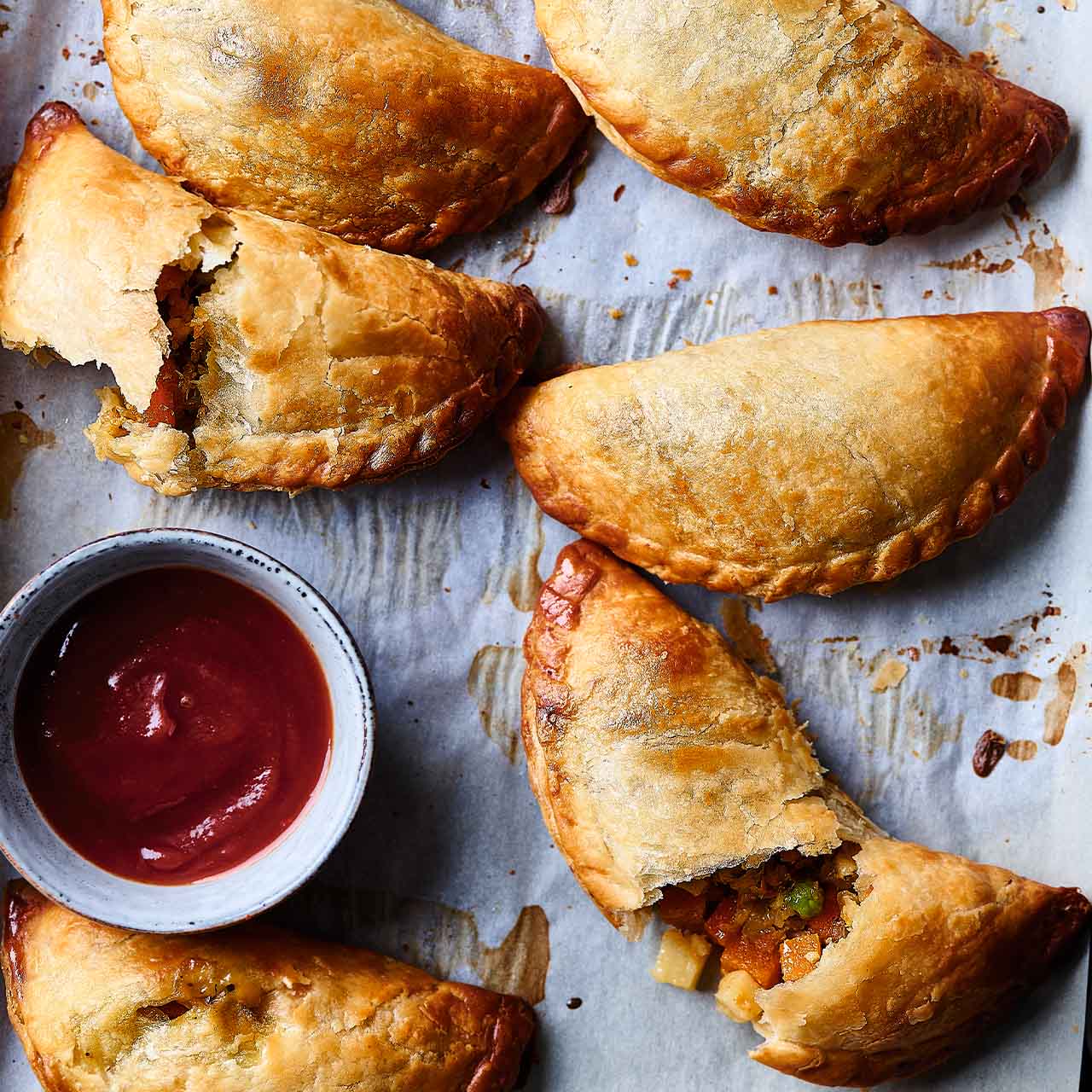 Cornish pasties filled with vegetables, one broken open to reveal the hearty filling, alongside a bowl of tomato sauce.