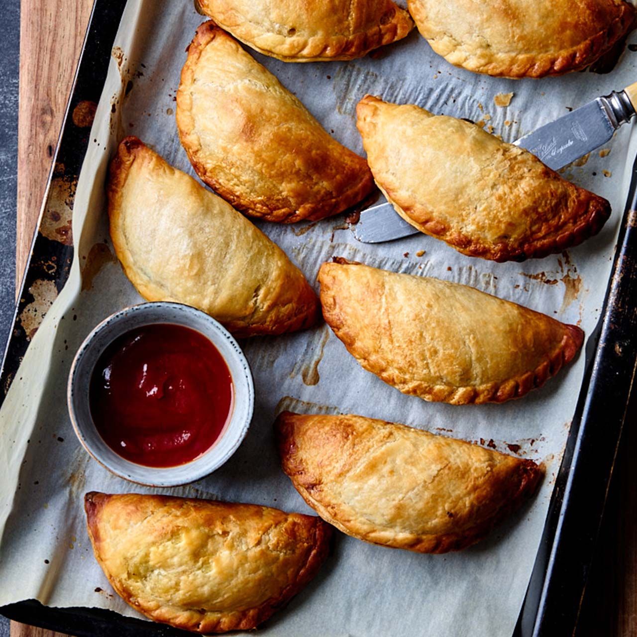 Freshly baked Cornish pasties arranged on a tray, with crisp golden pastry and a dipping bowl of tomato sauce.