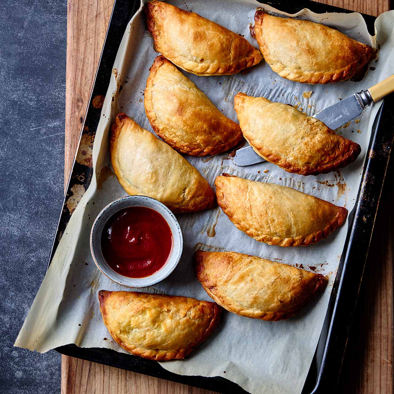 Golden Cornish pasties on a baking tray lined with parchment, served with a small bowl of tomato sauce for dipping.