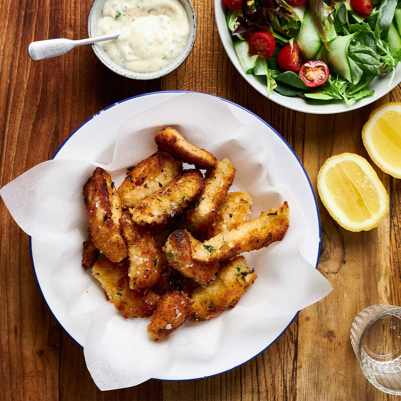 Overhead view of golden panko-crusted Monkfish Fish Fingers in a bowl, served with lemon wedges, salad, and dipping sauce.
