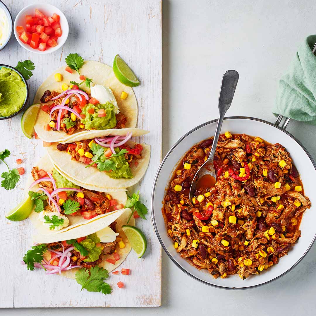Overhead view of tacos filled with Mexican shredded chicken, guacamole, pickled onions and coriander, with a bowl of chicken and corn on the side.