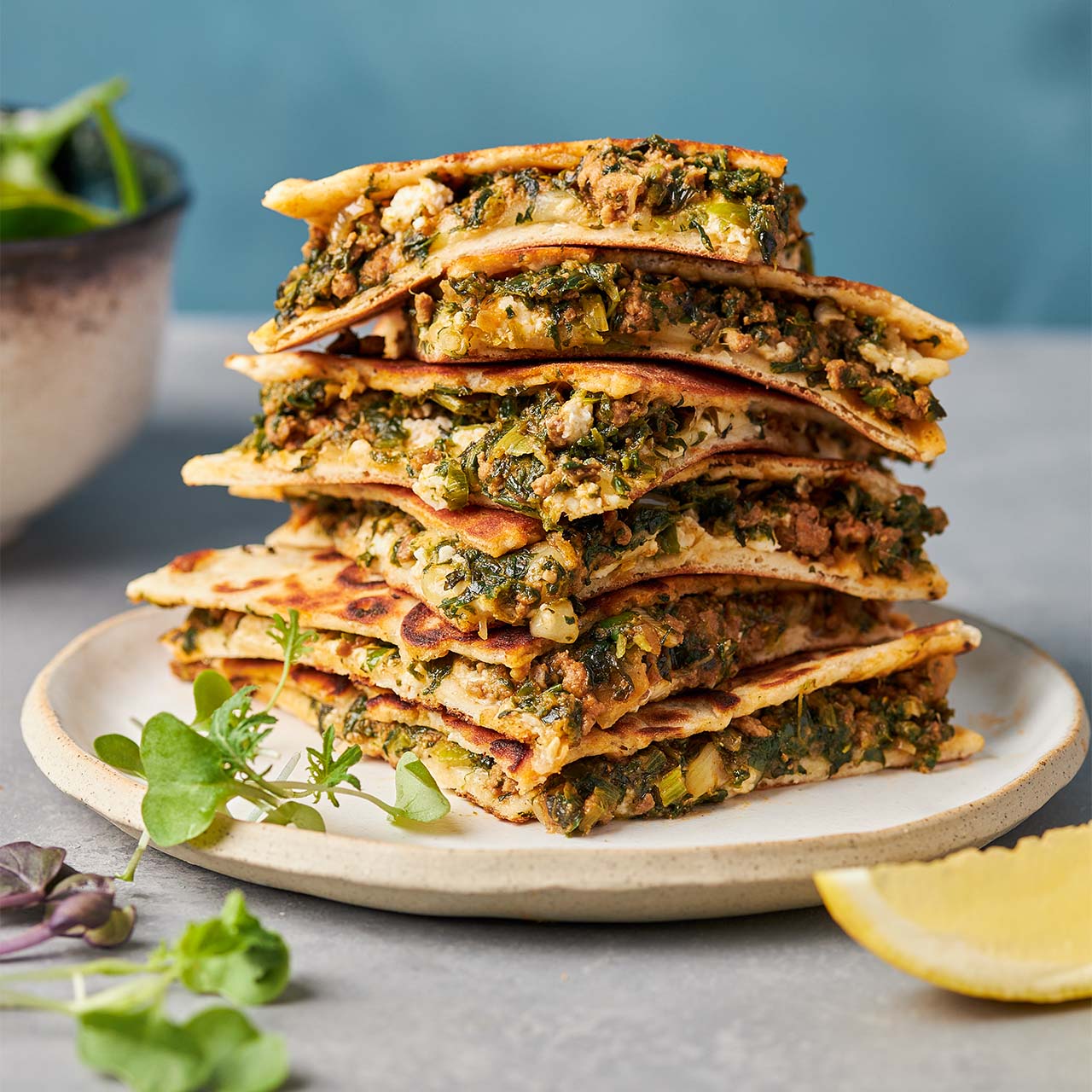 Stack of Middle Eastern lamb flat breads filled with spiced lamb, spinach, mint, and cheese, served on a plate with fresh greens and a lemon wedge.