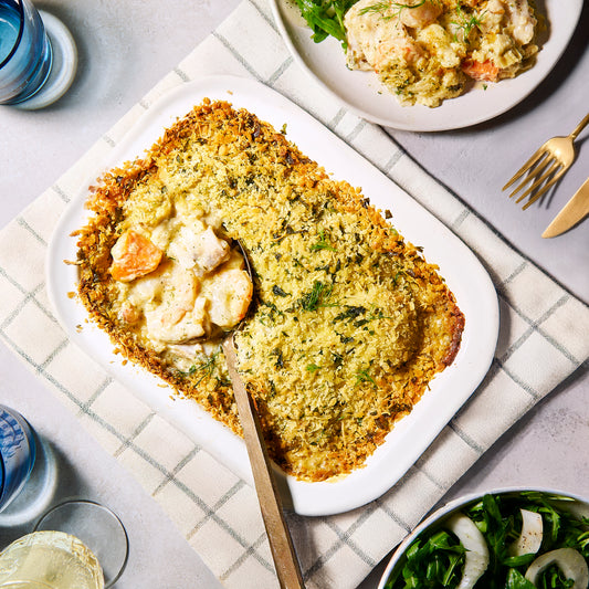 Overhead view of Prawn, Leek & Barramundi Gratin in a white baking dish with golden crumb topping, served with salad on the side.