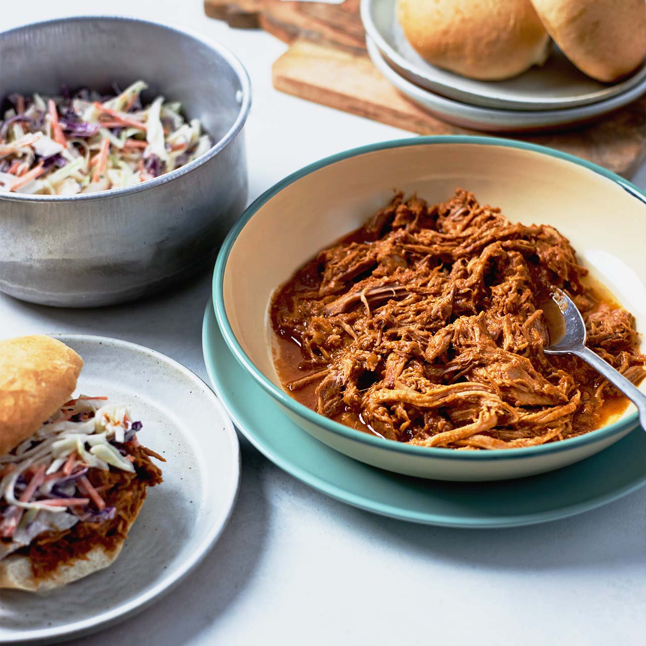 Pulled pork with BBQ sauce served in a bowl, alongside fresh coleslaw and bread rolls, ready for sandwiches or sliders.