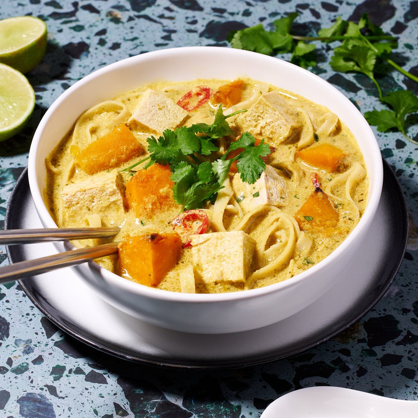 Pumpkin and tofu laksa in a white bowl, topped with coriander, served with lime wedges on the side.