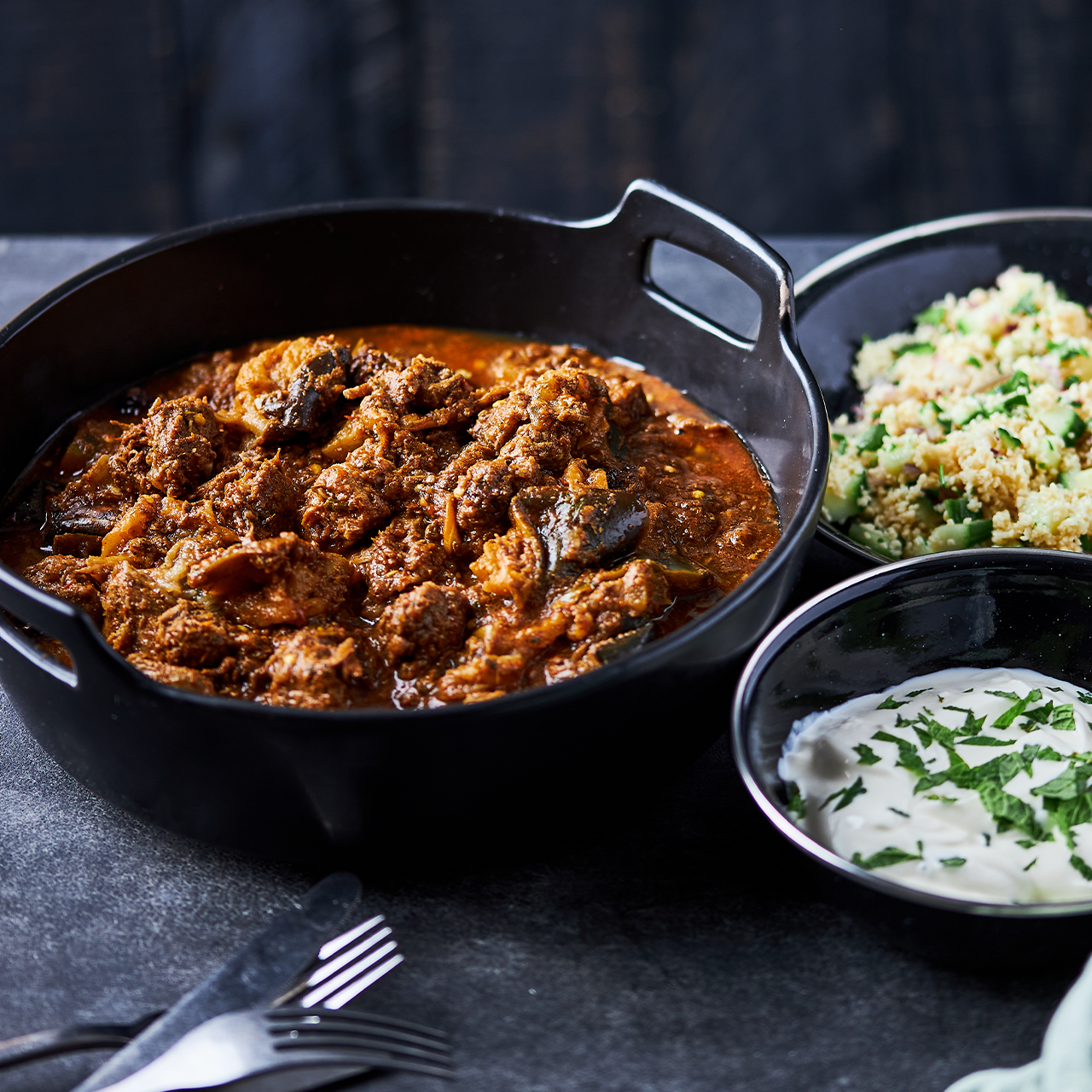 Slow-braised lamb with eggplant in a tomato and cinnamon sauce, served in a black pot with bowls of fluffy couscous and yoghurt on the side.