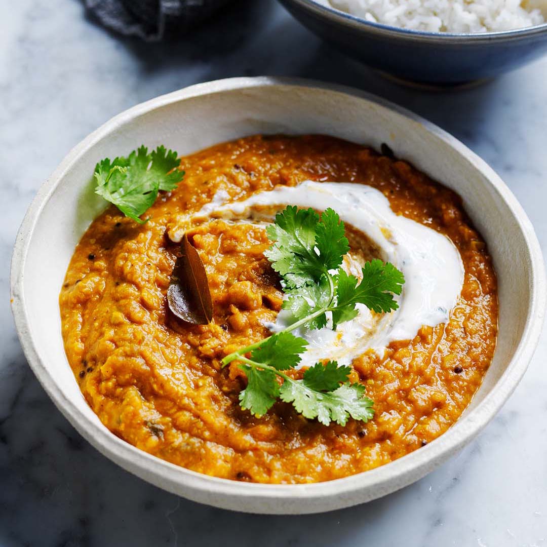Bowl of Tarka Dahl made with creamy spiced lentils, garnished with fresh coriander and yoghurt, served alongside steamed rice.