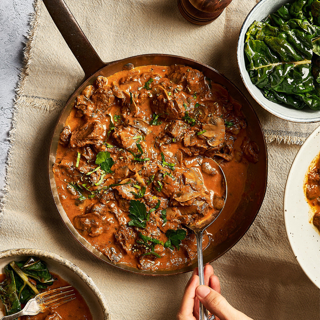 A skillet filled with slow-cooked beef stroganoff in a creamy mushroom sauce, garnished with fresh herbs, served alongside greens and sides