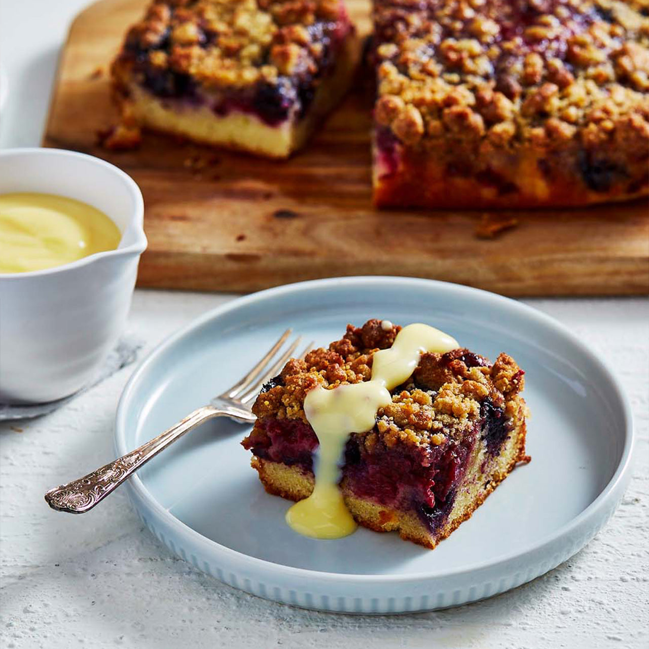 A slice of berry crumble cake on a plate, topped with custard, with the full cake in the background on a wooden board.