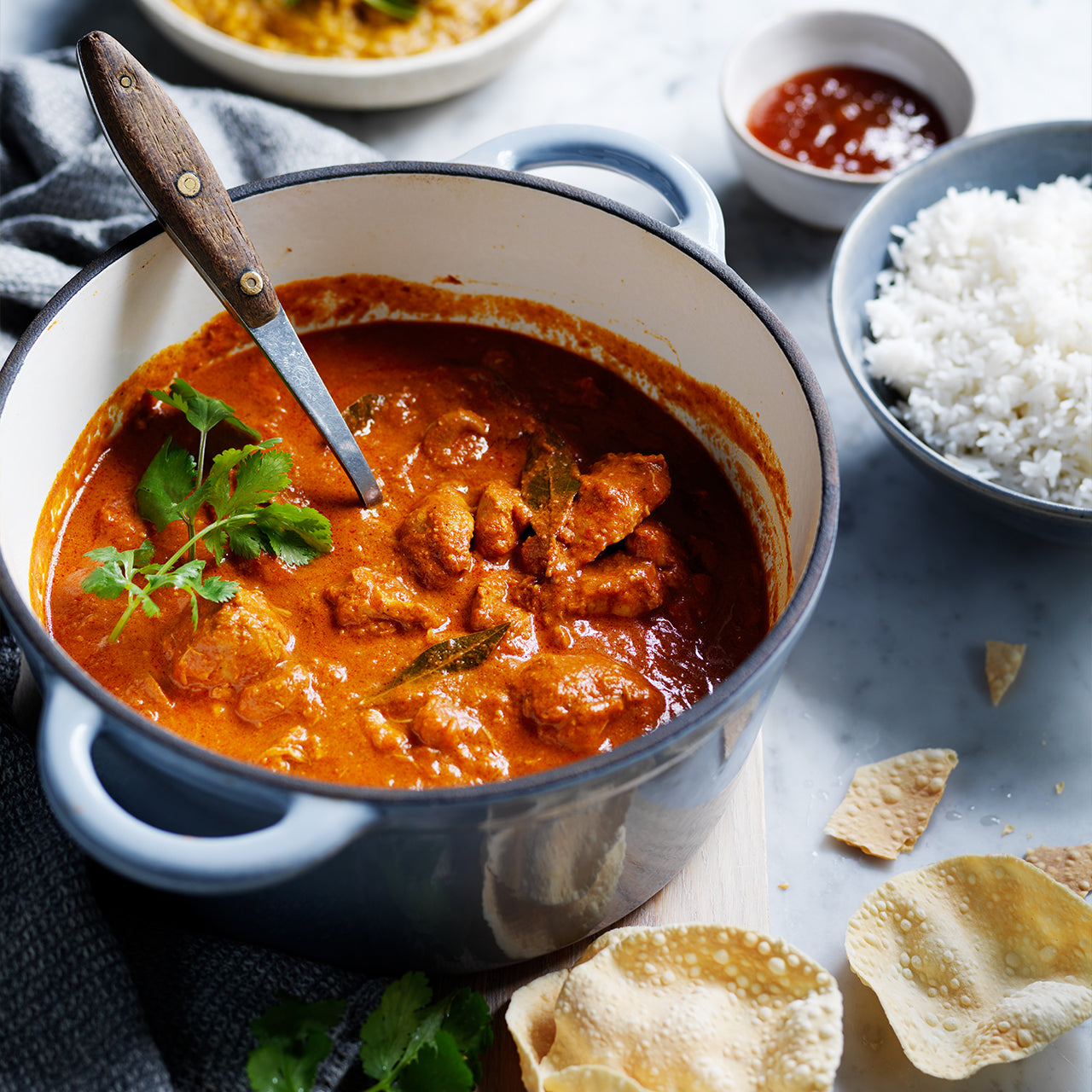 Butter Chicken in a pot with creamy tomato sauce, served with rice and poppadoms.