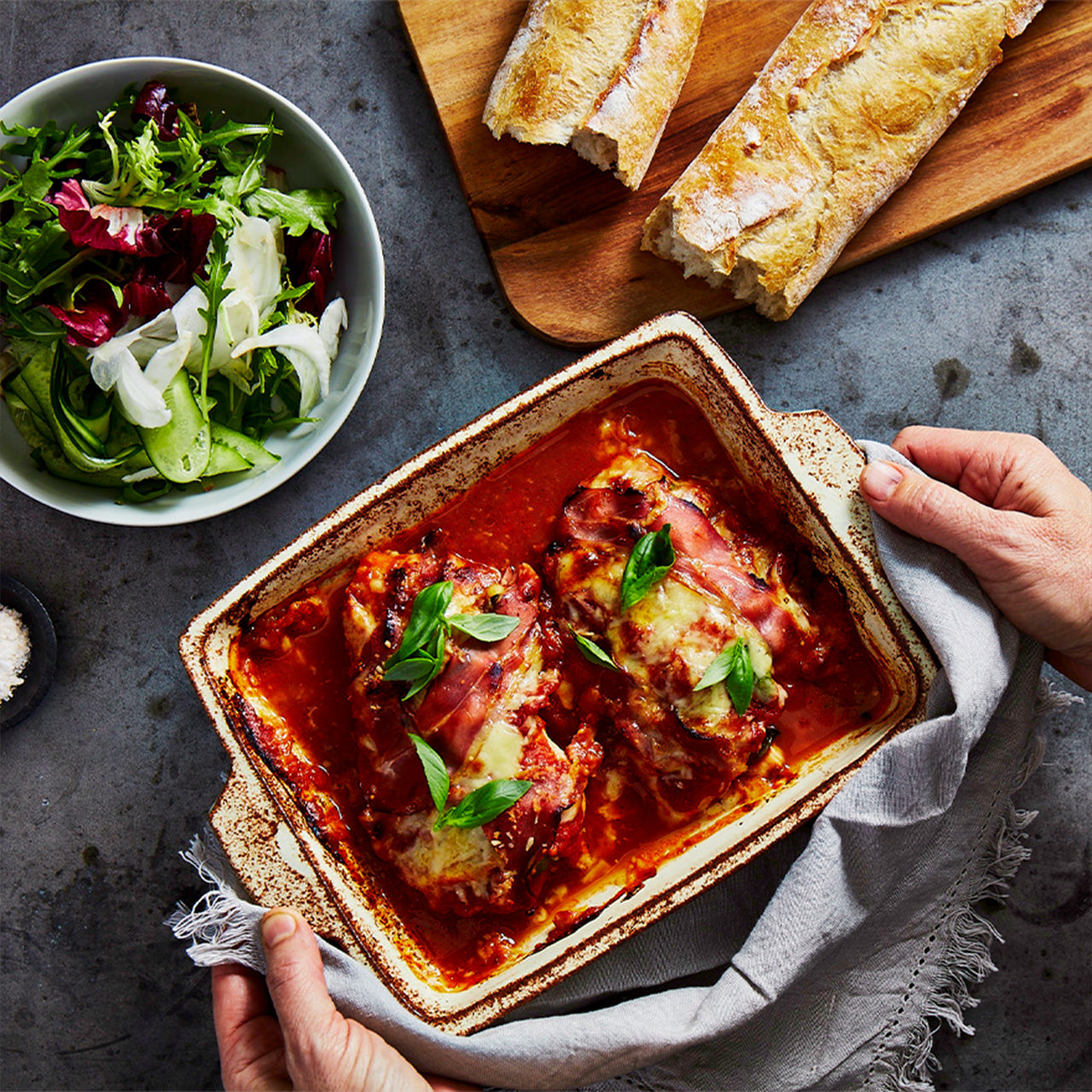 Chicken Parmigiana baked with tomato sauce, prosciutto, mozzarella & Parmesan, garnished with basil, served in a tray with salad & bread.