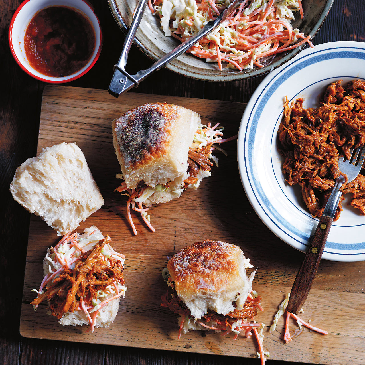 Pulled pork with BBQ sauce in crusty bread rolls topped with coleslaw, served on a wooden board with extra pork and slaw.