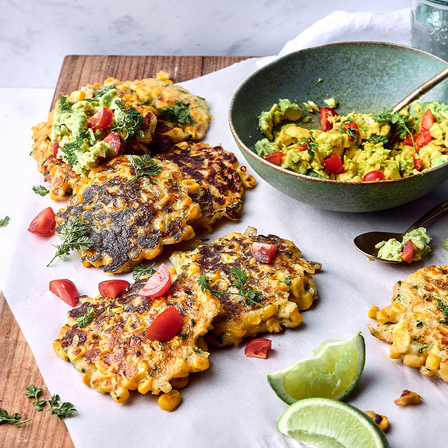 Overhead view of Sweetcorn & Haloumi Fritters topped with cherry tomatoes and herbs, served with smashed avocado and lime.