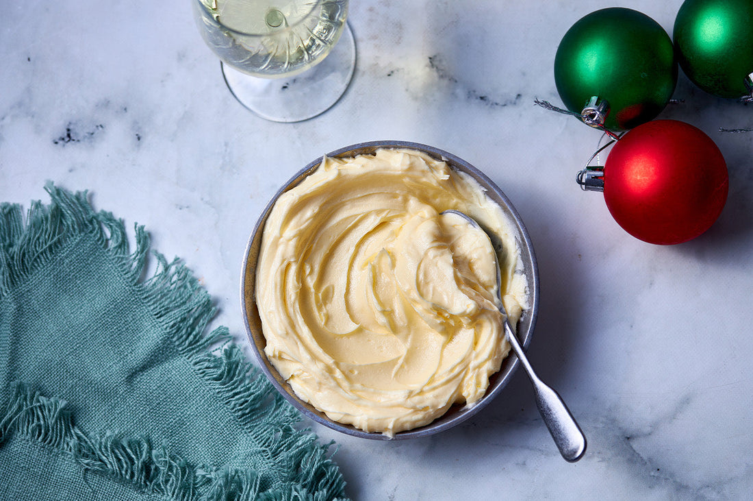 Bowl of creamy brandy butter with a spoon, set on marble with festive decorations and a green cloth.