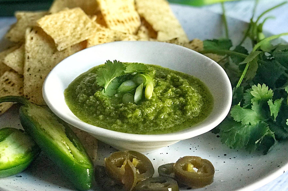 Bowl of coriander-jalapeño salsa with tortilla chips, jalapeños, and herbs on a plate.