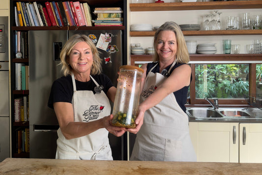 Two women holding a jar of smashed cucumber salad in a bright kitchen.