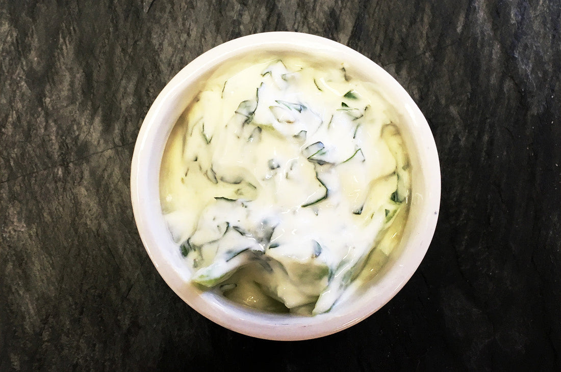 Bowl of cucumber raita with yoghurt and fresh herbs on a dark surface.