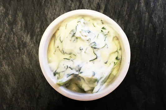 Bowl of cucumber raita with yoghurt and fresh herbs on a dark surface.