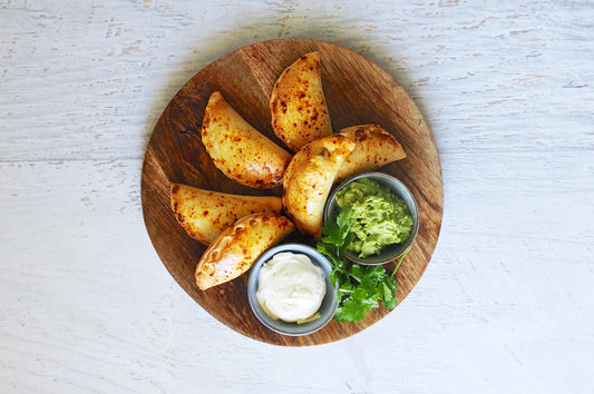 Wooden board with golden baked empanadas served alongside guacamole, sour cream, and fresh coriander.