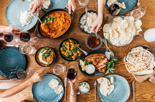 Overhead view of a curry feast with naan, rice, chutneys, and shared dishes, surrounded by hands serving at a dinner table.