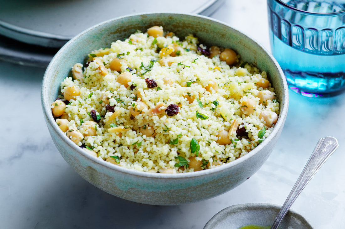 Bowl of couscous with chickpeas, herbs, and dried fruit on a marble table.