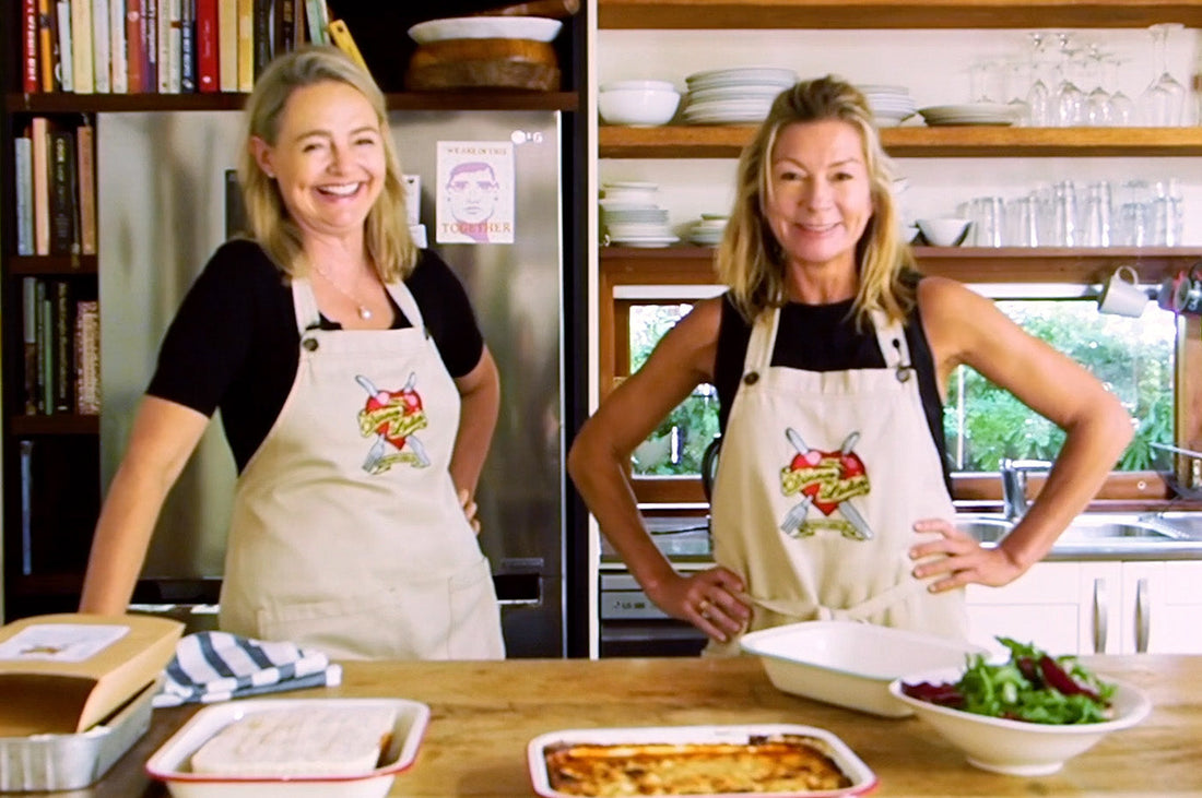 The Dinner Ladies founders in aprons smiling in a kitchen with a prepared meal on the counter.