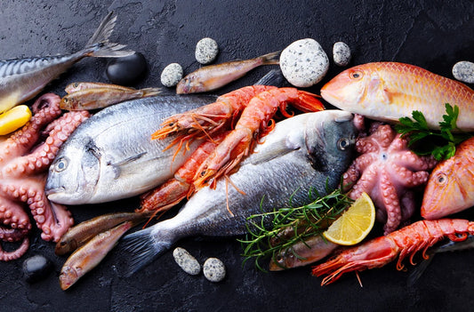 Fresh seafood display with fish, prawns, octopus, herbs, and lemon on a dark background.