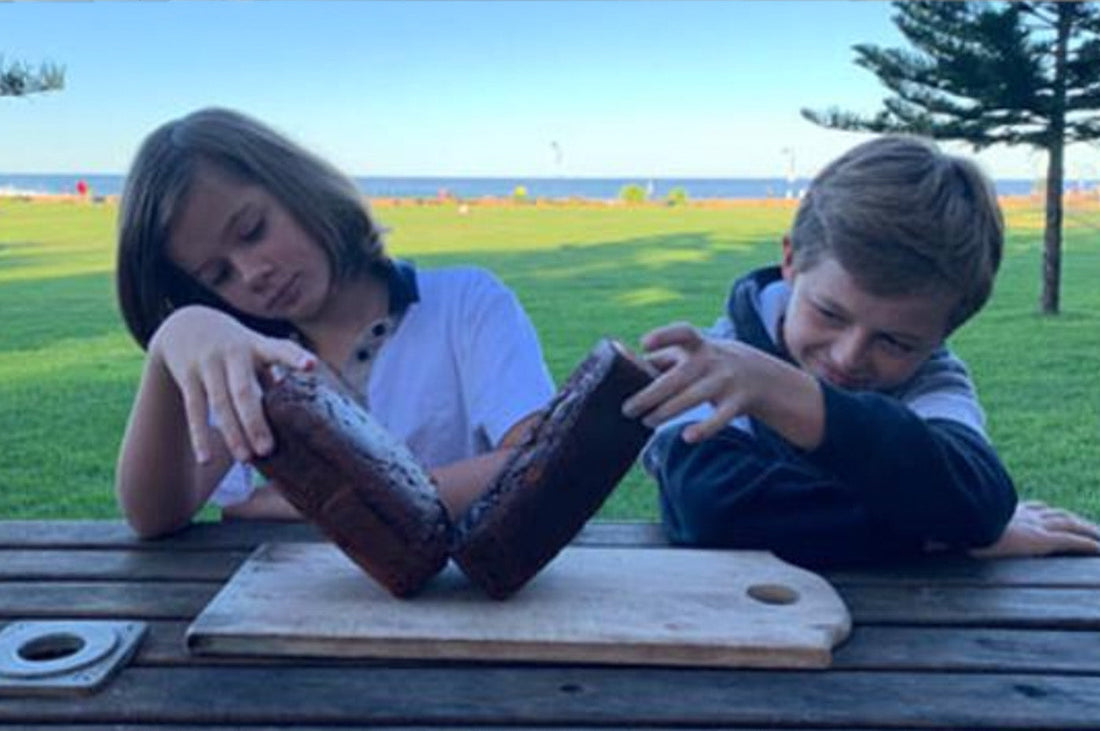 Two kids playfully balancing loaves of zucchini bread on a wooden board outdoors.
