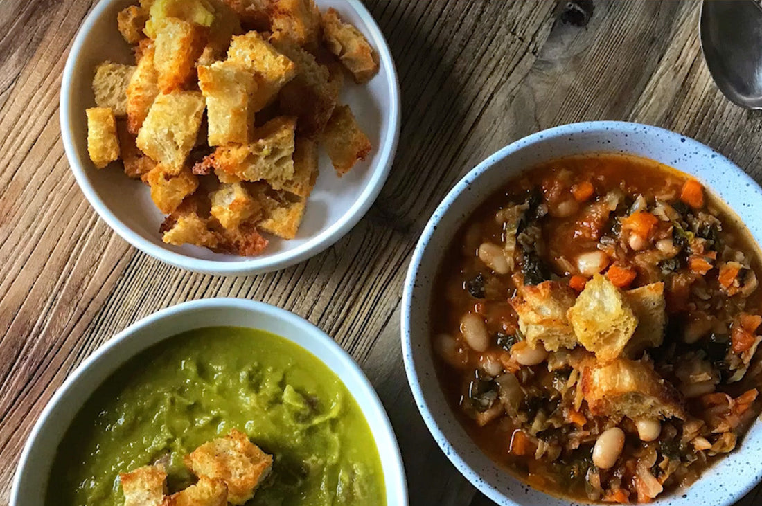 Bowls of Parmesan croutons, green soup, and bean stew on a wooden table.