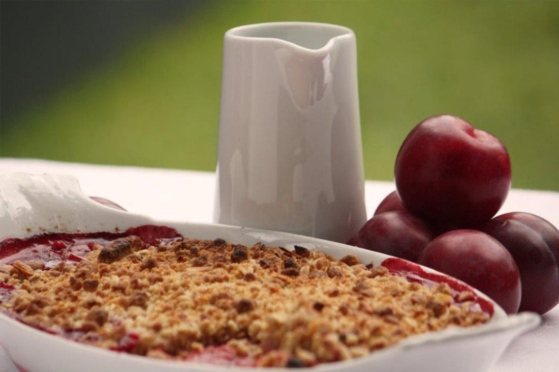 Plum and raspberry granola crumble in a white dish beside fresh plums and a jug.