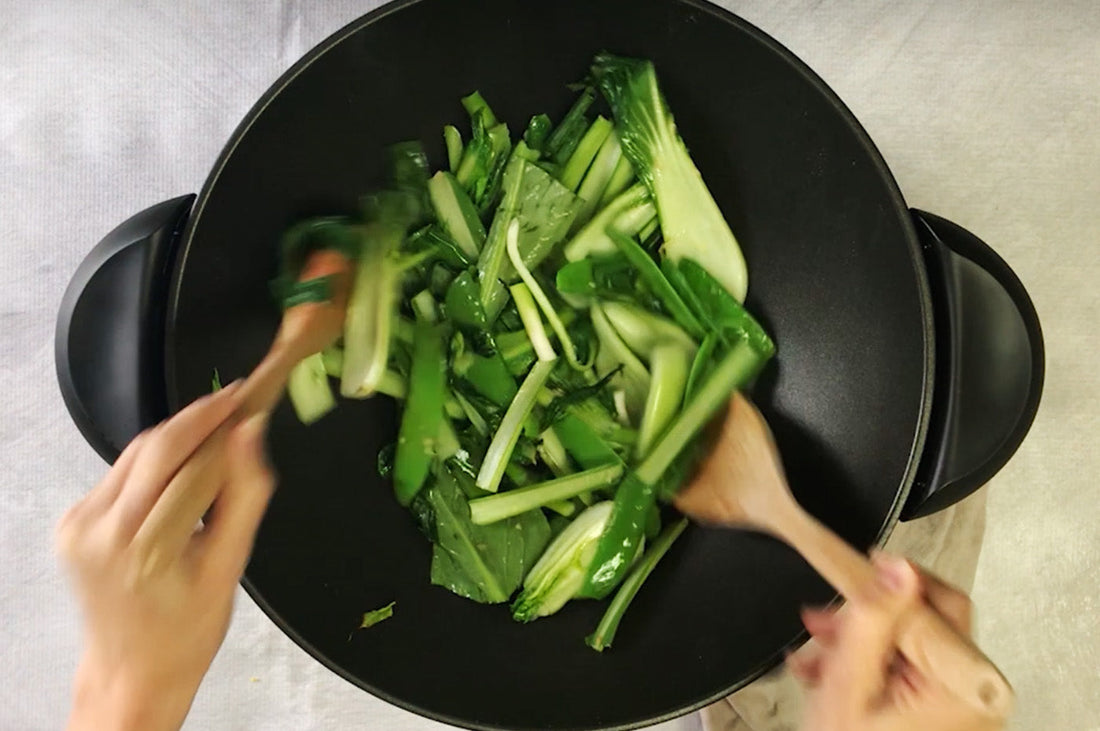 Hands stir-frying Asian greens in a black wok with wooden utensils.