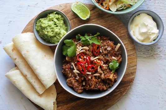 Bowl of pork picadillo with tortillas, guacamole, lime, and sour cream on a board.