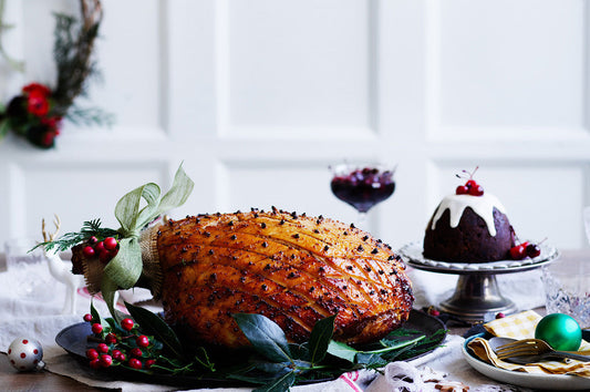 Glazed Christmas ham decorated with cloves, styled on a festive table with pudding and holiday decorations.