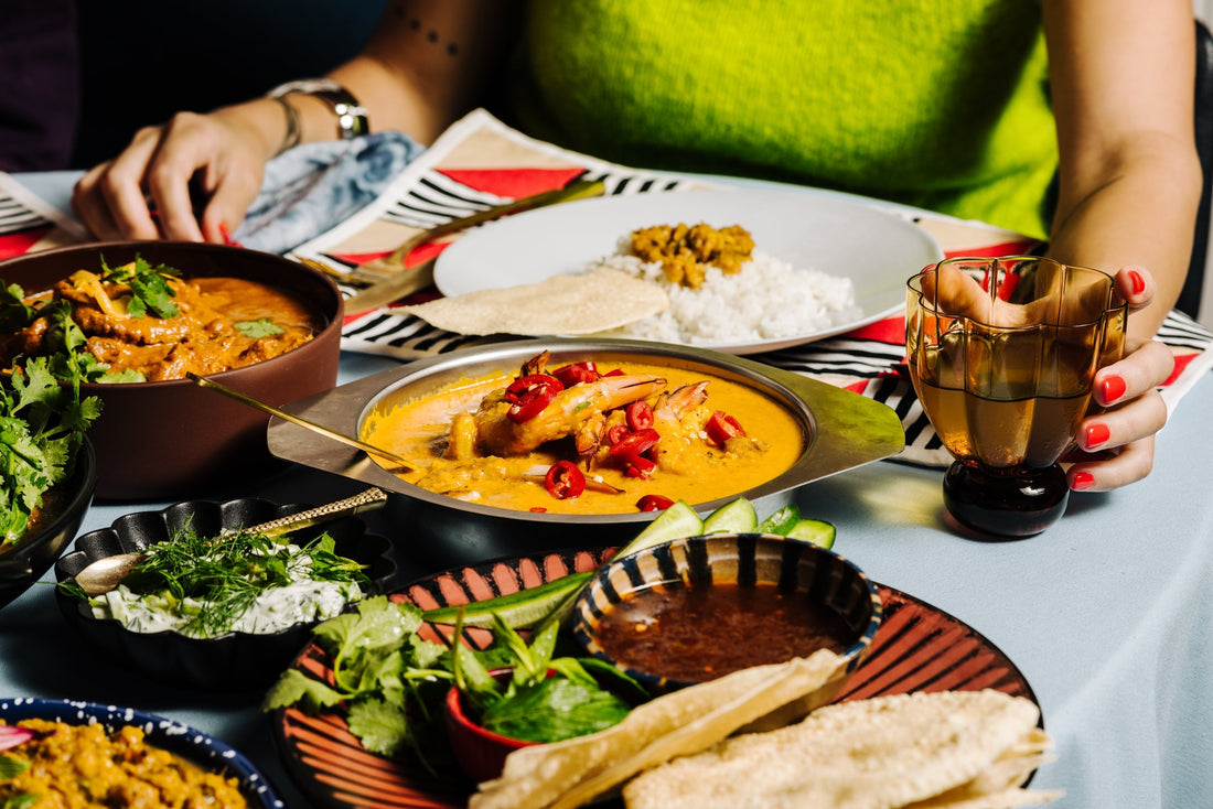 Table of Dinner Ladies curries and sides with a person reaching for a drink.