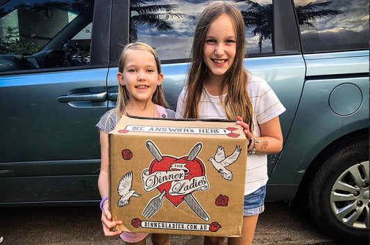 Two smiling children holding a Dinner Ladies delivery box filled with nourishing meals, symbolising giving back to the community.