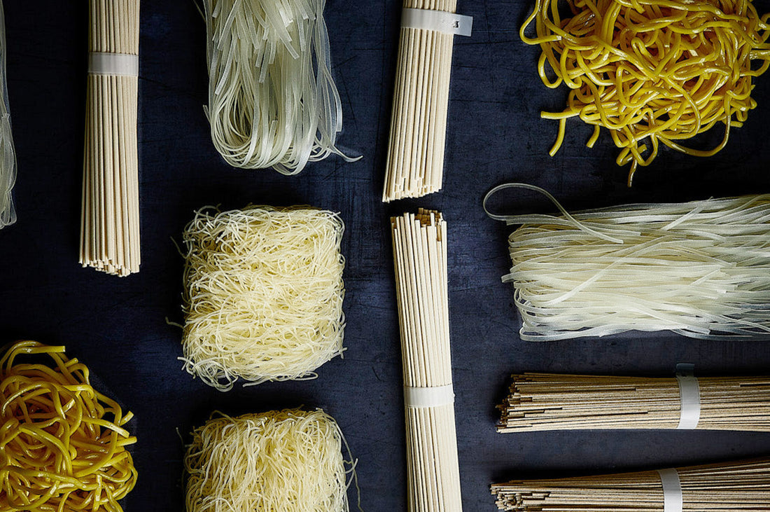 Assorted bundles of uncooked noodles and pasta, including rice noodles, egg noodles, and wheat noodles, arranged neatly on a dark background.