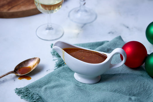 White gravy boat filled with orange, maple syrup and thyme glaze for Christmas ham, set on a green cloth with festive baubles.