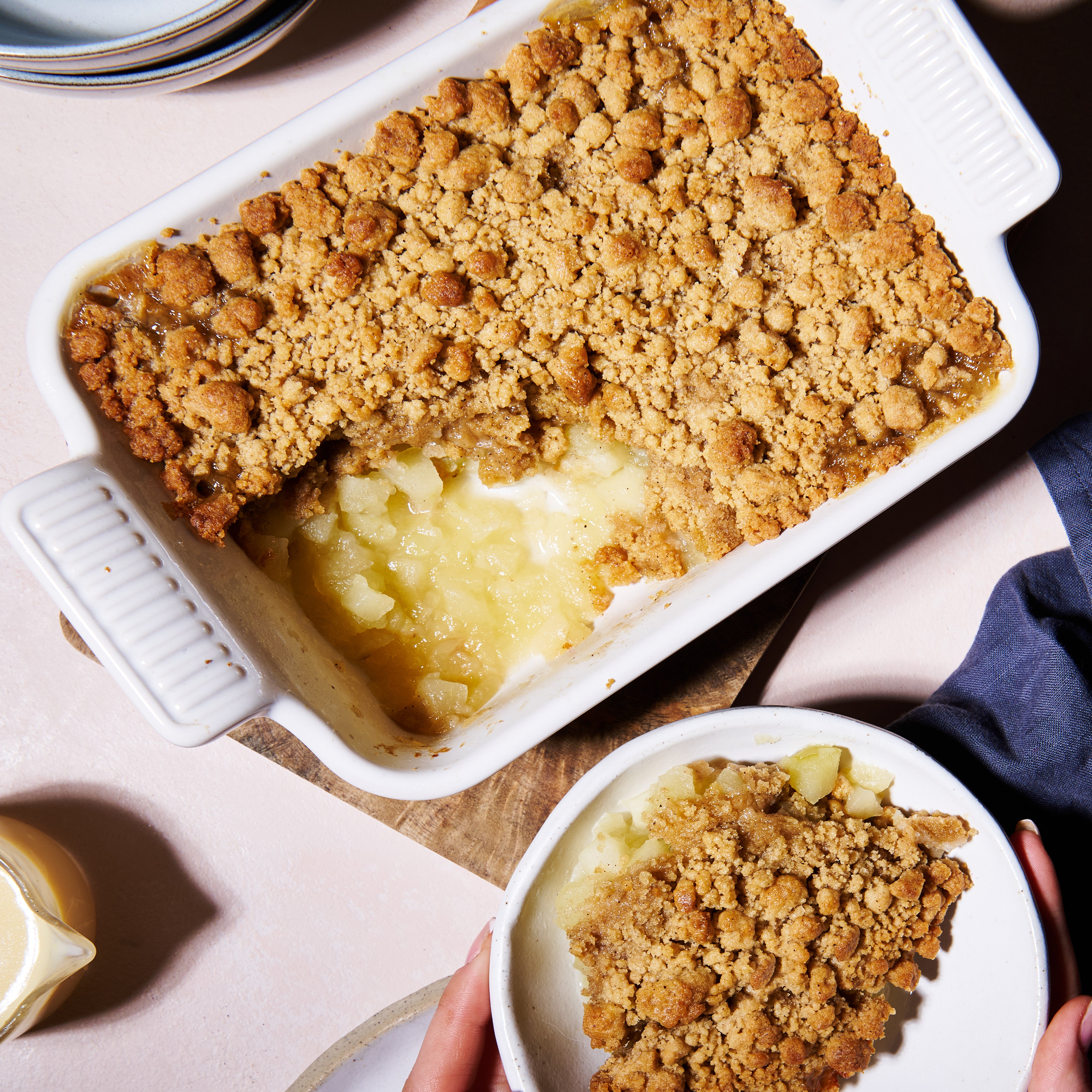 apple and gingerbread crumble, served in a white baking dish, alongsidee a portion plated. Golden brown crumble with stewed apple visible.