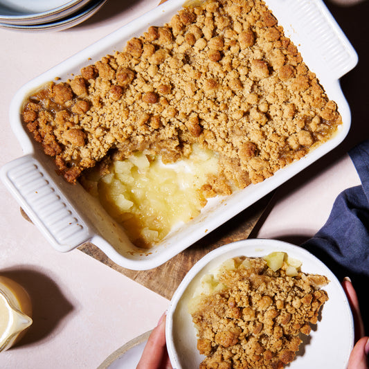 apple and gingerbread crumble, served in a white baking dish, alongsidee a portion plated. Golden brown crumble with stewed apple visible.