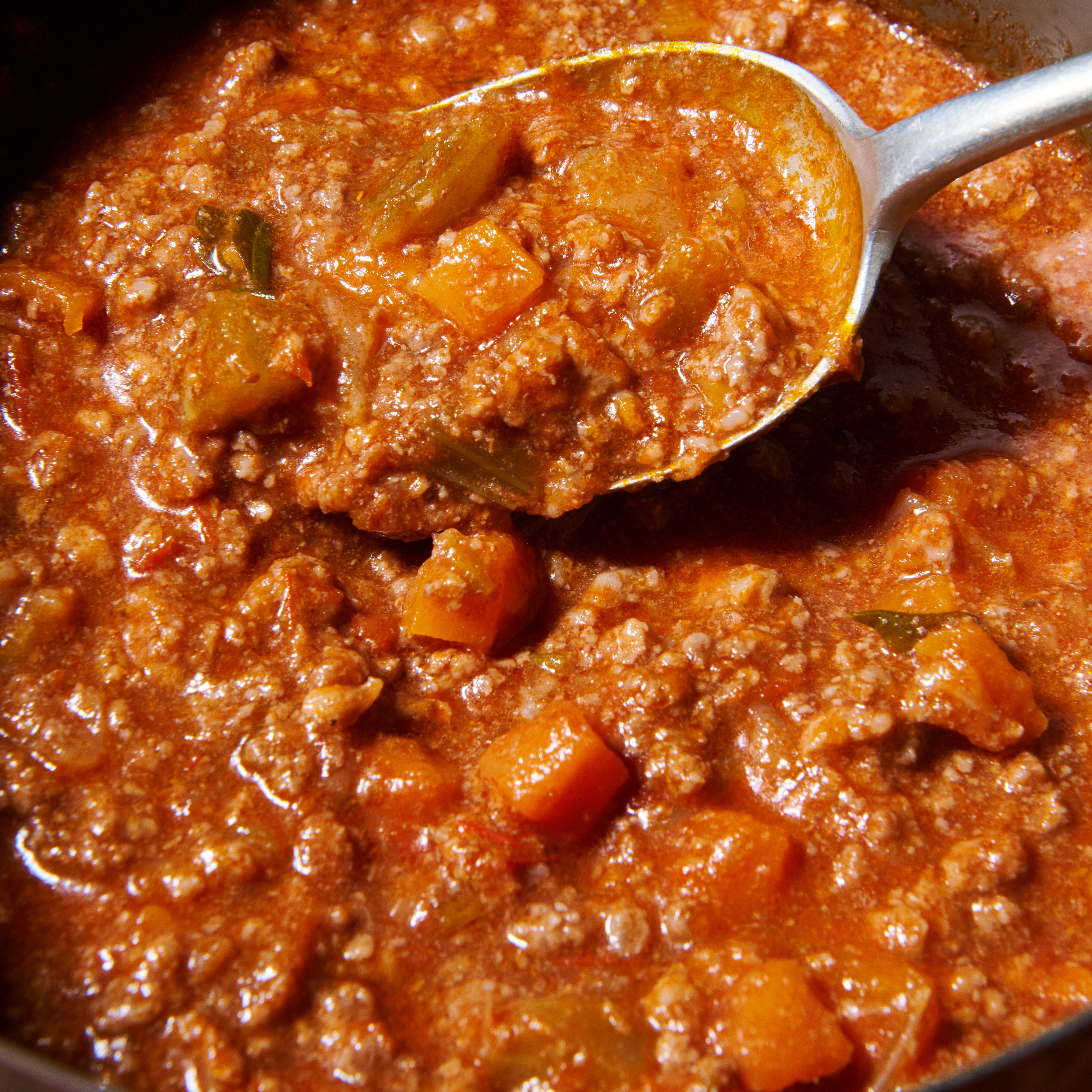 Close-up of a spoon lifting beef bolognese, showing minced beef, carrot and celery in a rich tomato sauce