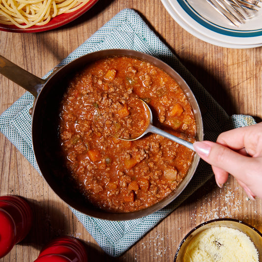 Beef bolognese in a rich tomato sauce with carrot and celery, in a pot with a spoon, served alongside spaghetti